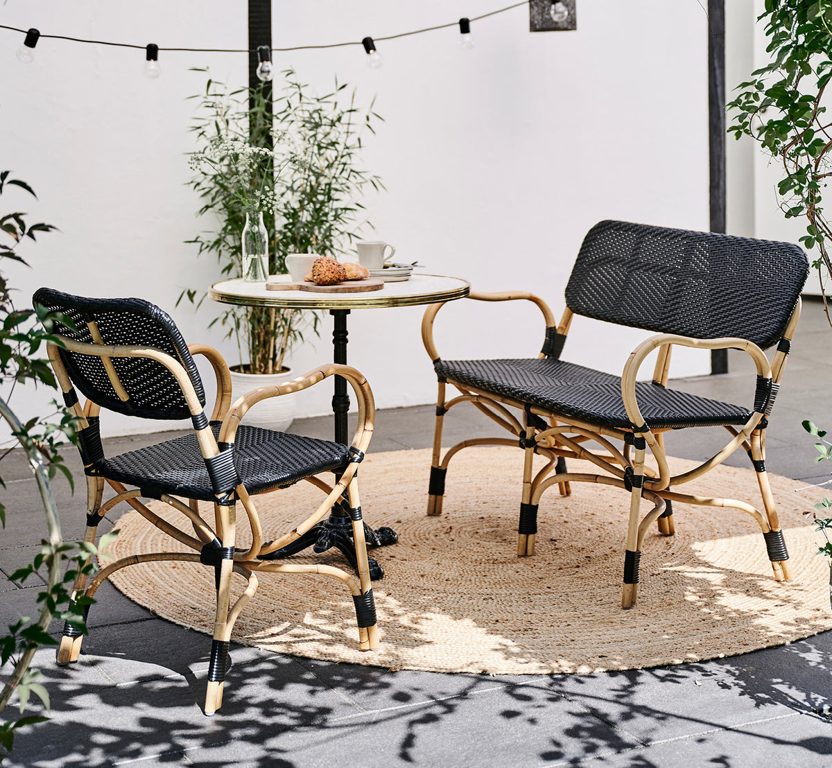 Esta es una imagen de estilo de vida de Bistro Bench en skin on Rattan frame, en un ambiente de café al aire libre con una mesa de bistró sobre.