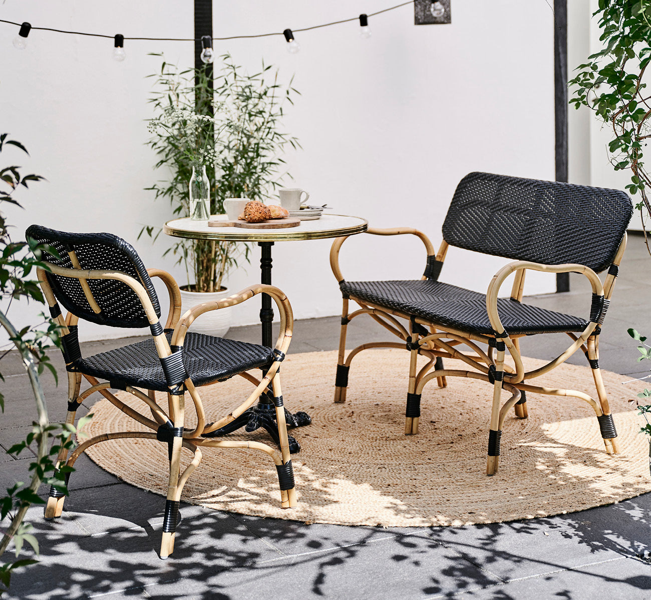 Esta es una imagen de estilo de vida de Bistro Bench en skin on Rattan frame, en un ambiente de café al aire libre con una mesa de bistró sobre.