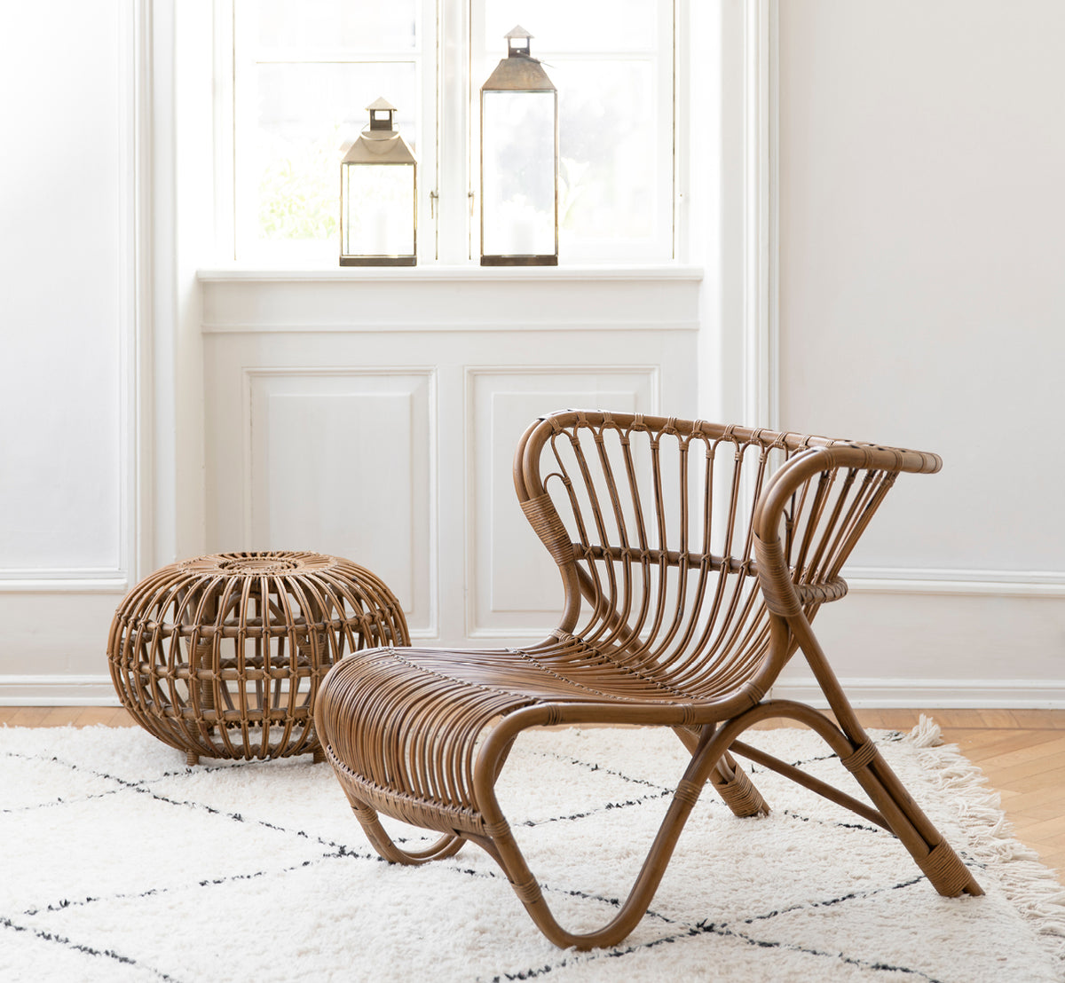 Esta es una imagen de estilo de vida de Ottoman Small en polished antique rattan. Colocado en una habitación con papel pintado ornamentado y una estatua clásica.