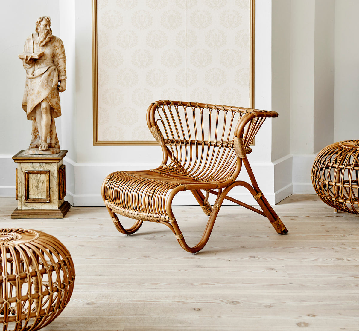 Esta es una imagen de estilo de vida de Ottoman Small en polished antique rattan. Colocado en una habitación con papel pintado ornamentado y una estatua clásica.