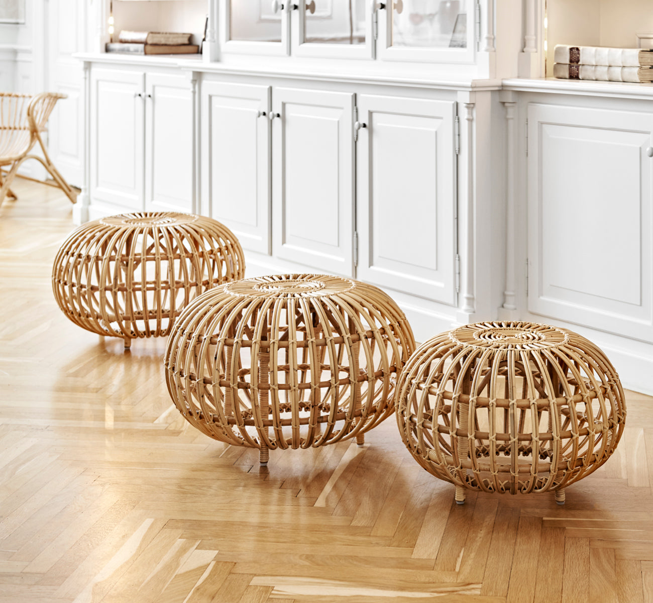 Esta es una imagen de estilo de vida de Ottoman Small en natural skin on rattan. Agrupados en un interior clásico con paneles y un reloj antiguo.