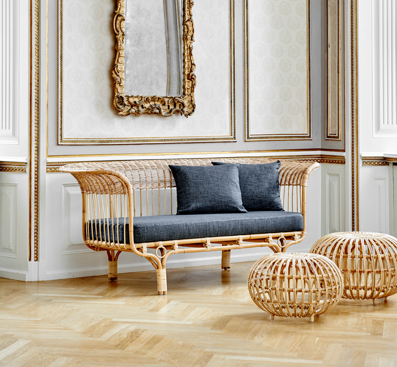 Esta es una imagen de estilo de vida de Ottoman Small en natural skin on rattan. Agrupados en un interior clásico con paneles y un reloj antiguo.