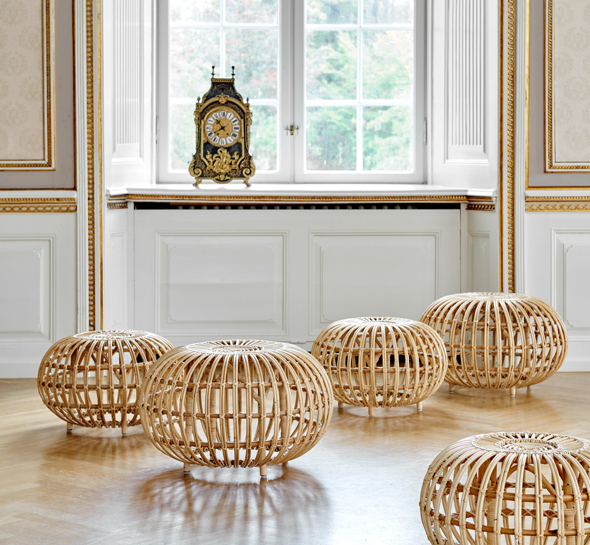 Esta es una imagen de estilo de vida de Ottoman Large en natural skin on rattan. Agrupados en un interior clásico con paneles y un reloj dorado.