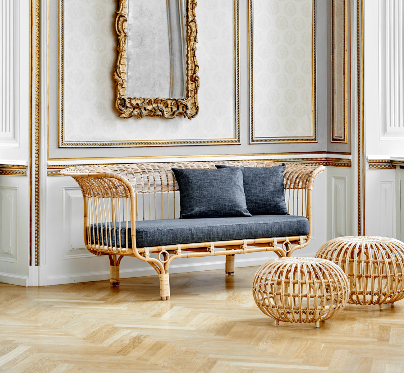 Esta es una imagen de estilo de vida de Ottoman Large en natural skin on rattan. Agrupados en un interior clásico con paneles y un reloj dorado.