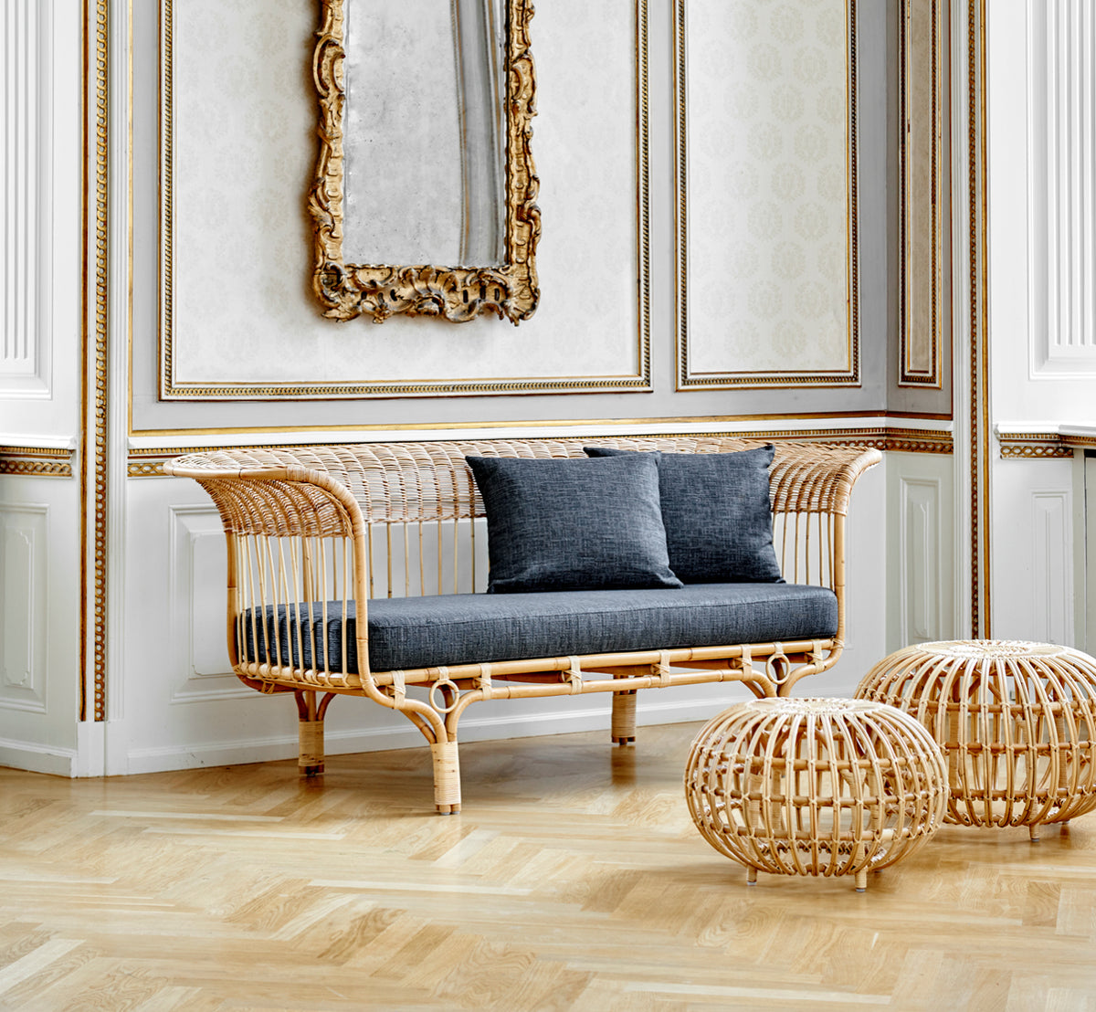 Esta es una imagen de estilo de vida de Ottoman Large en natural skin on rattan. Agrupados en un interior clásico con paneles y un reloj dorado.