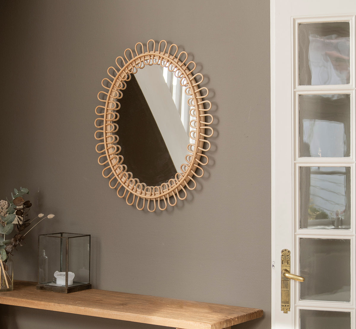 Esta es una imagen de estilo de vida de Luella espejo en polished natural rattan. Colgado en una pared gris sobre una estantería de madera con eucalipto y decoración de cristal.