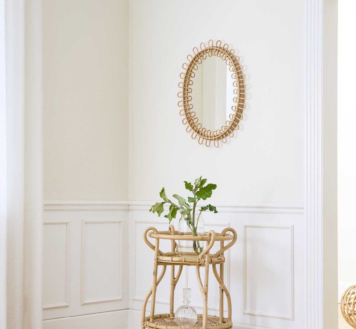 Esta es una imagen de estilo de vida de Luella espejo en polished natural rattan. Montado en una pared con paneles blancos sobre un carrito bar de ratán con plantas.