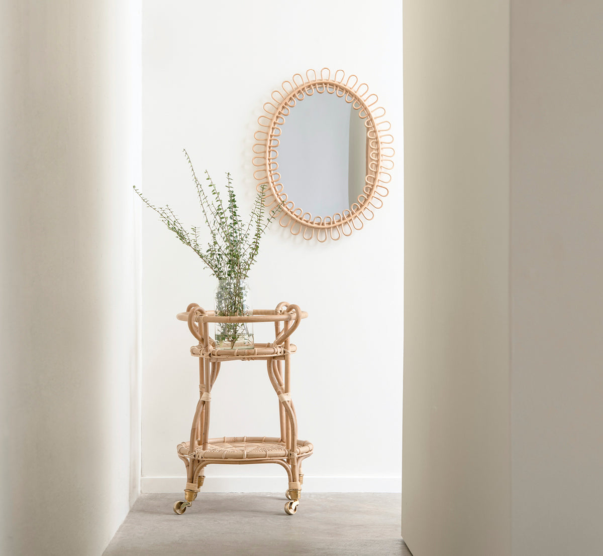Esta es una imagen de estilo de vida de Luella espejo en polished natural rattan. En una luminosa alcoba de pasillo sobre un carrito bar de ratán con plantas frescas.