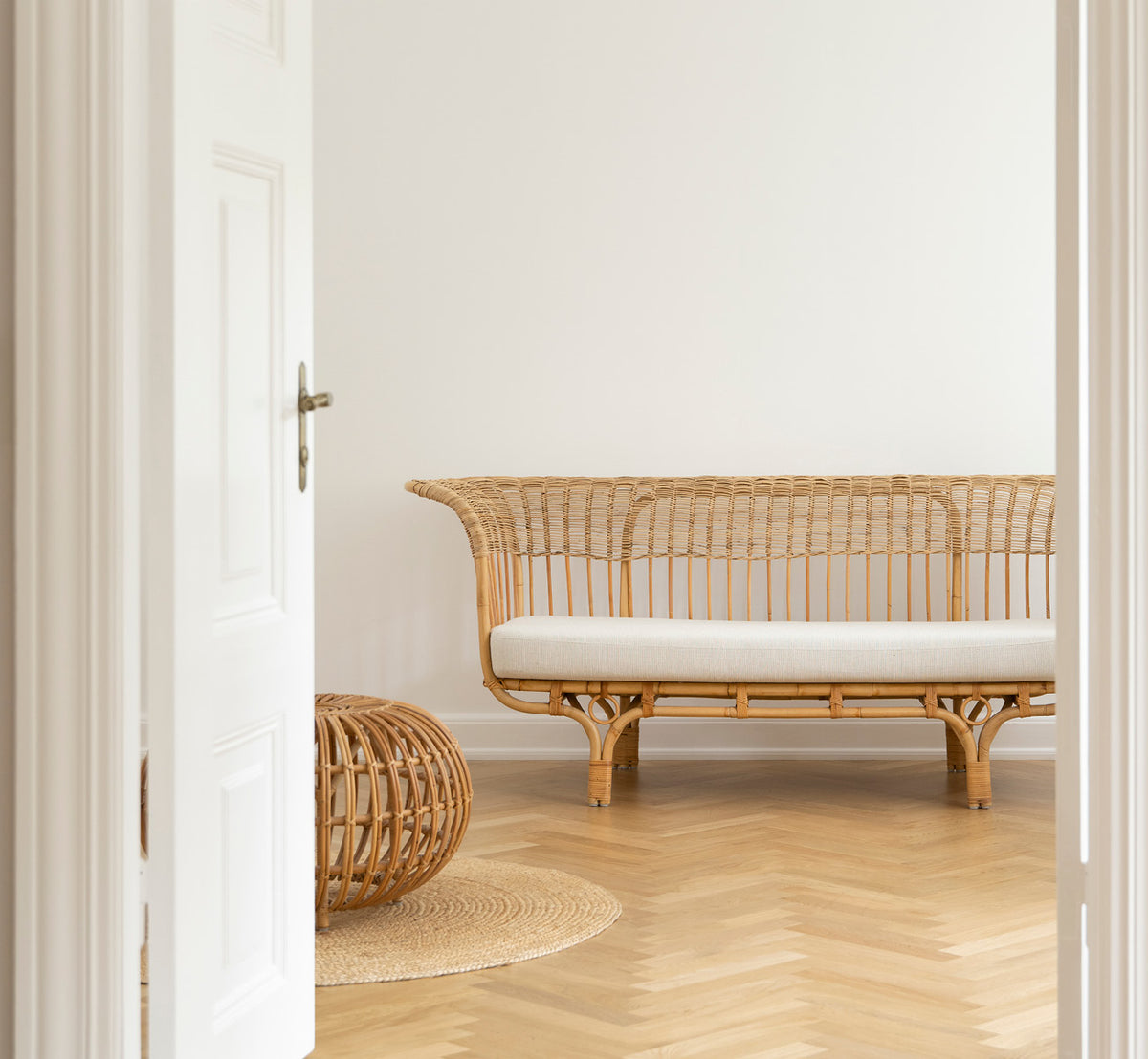 Esta es una imagen de estilo de vida de Belladonna sofá en polished natural rattan, en un luminoso interior escandinavo visto a través de un marco de.