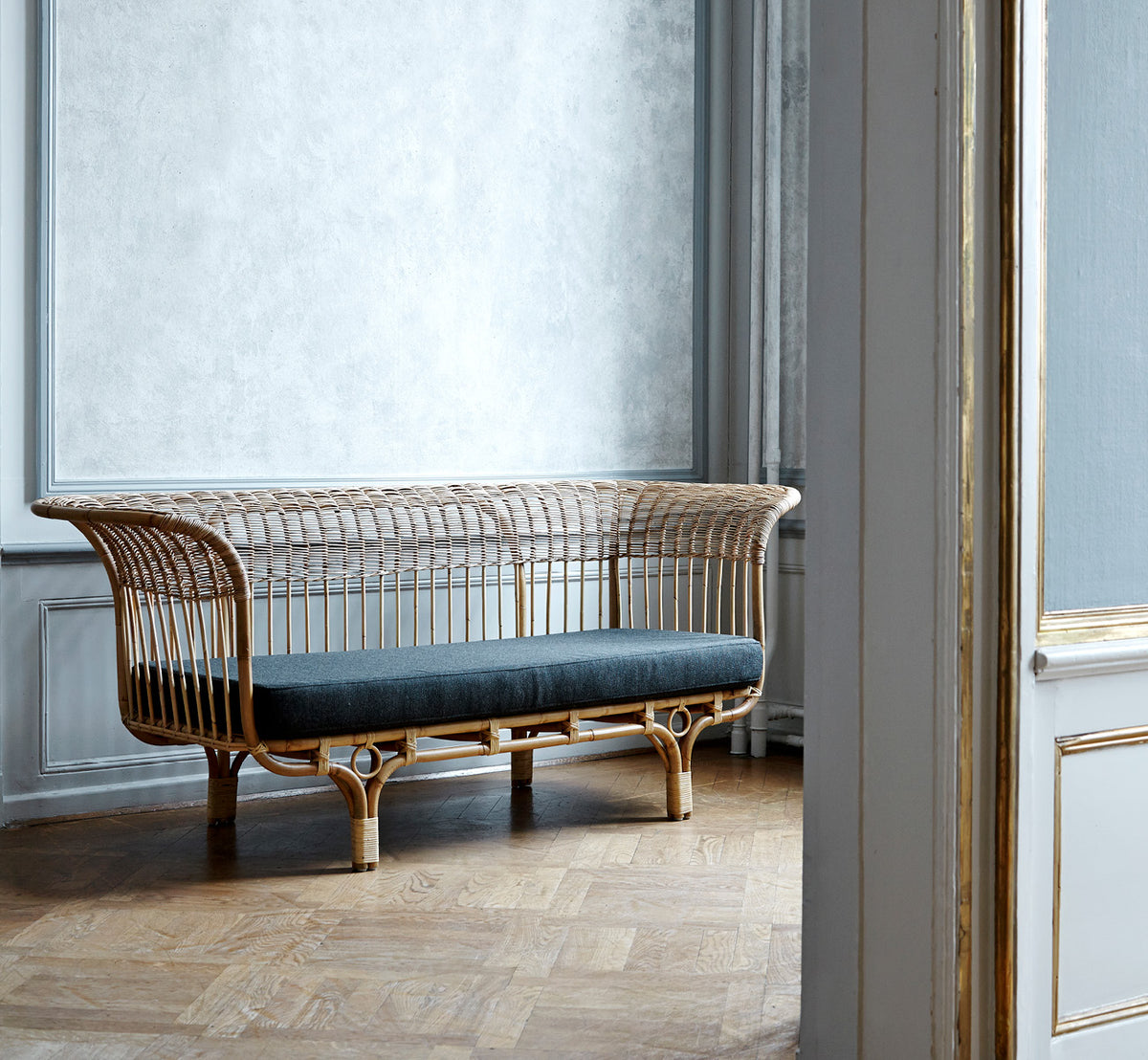 Esta es una imagen de estilo de vida de Belladonna sofá en polished natural rattan, en un luminoso interior escandinavo visto a través de un marco de.