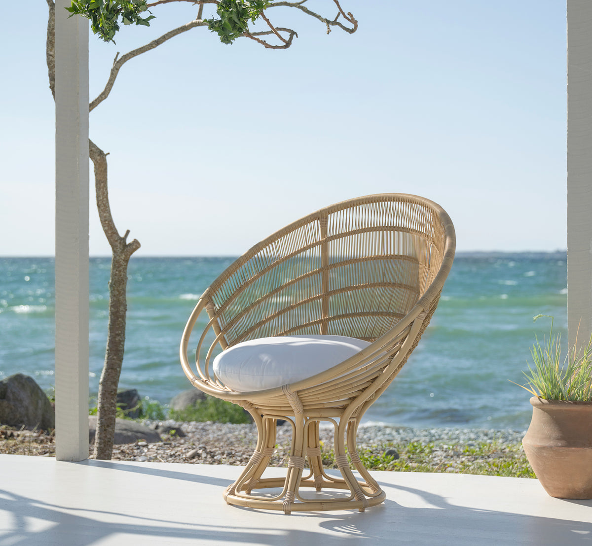 Esta es una imagen de estilo de vida de Luna sillón lounge exterior en natural aluminium artfibre. En una terraza blanca junto al mar con vista al océano y un árbol.