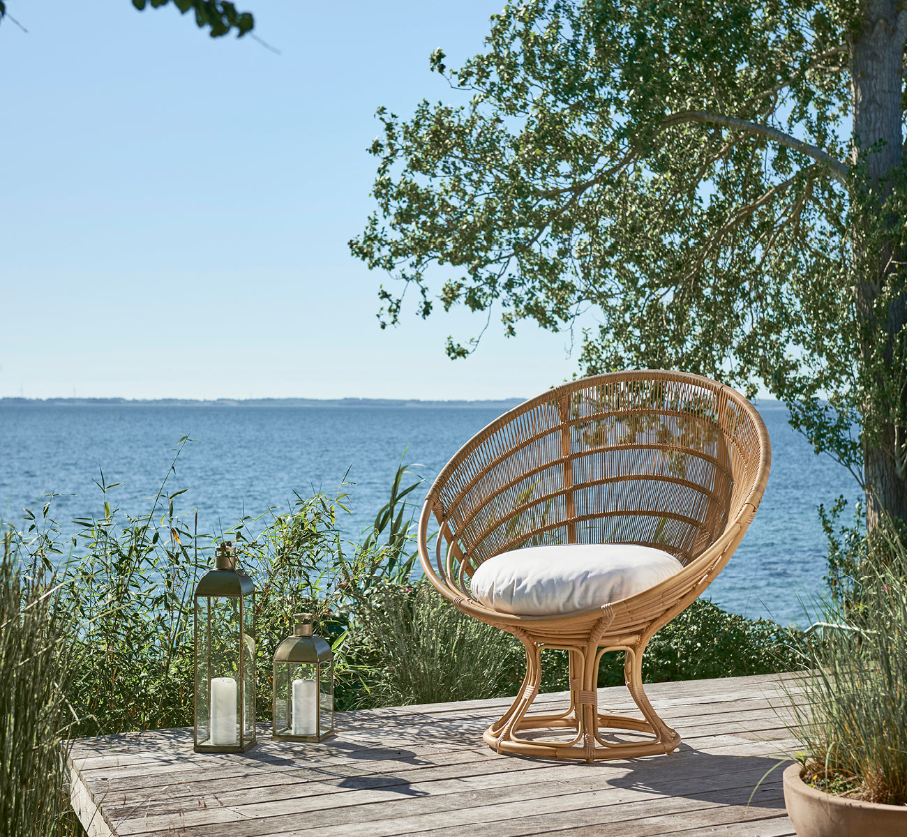 Esta es una imagen de estilo de vida de Luna sillón lounge exterior en natural aluminium artfibre. En una terraza de madera junto al mar rodeada de exuberante vegetación.