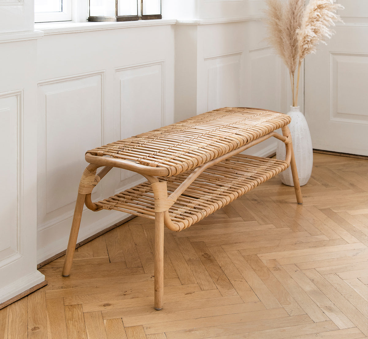 Esta es una imagen de estilo de vida de Machiya banco en natural skin on rattan. Pasillo con paneles blancos, suelo en espiga y jarrón con hierba de las pampas.