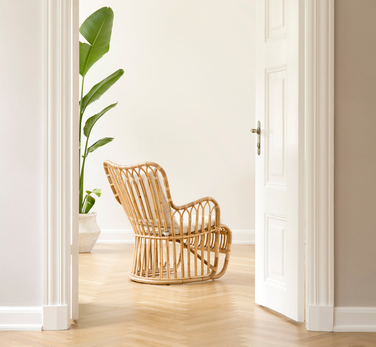 Esta es una imagen de estilo de vida de Tulip sillón lounge en polished natural rattan. Combinada con un sofá a juego en una sala luminosa con paneles y puerta.