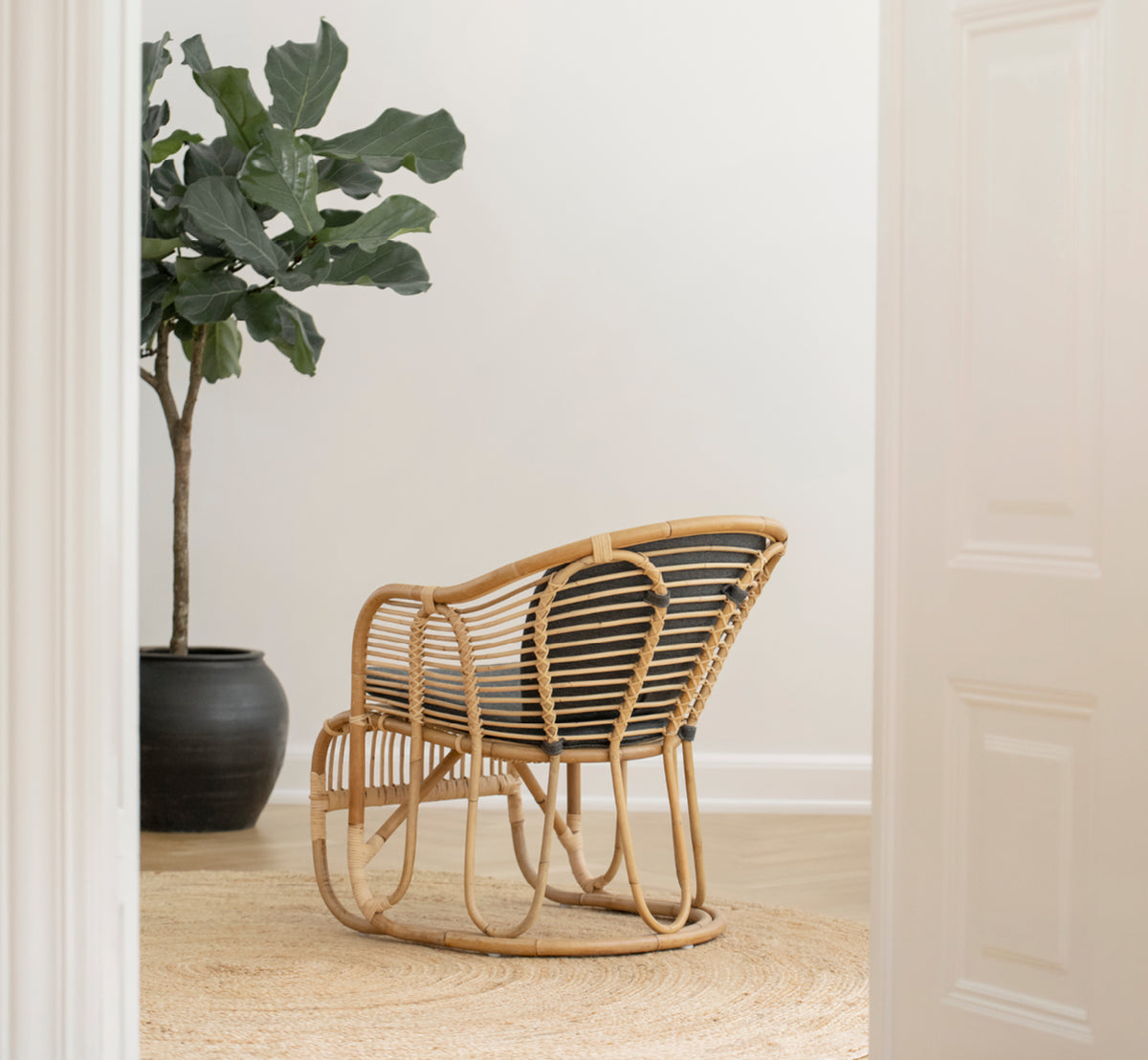 Esta es una imagen de estilo de vida de Swing sillón lounge en polished natural rattan. De pie sola junto a una planta frondosa en un suelo de parqué en espiga.