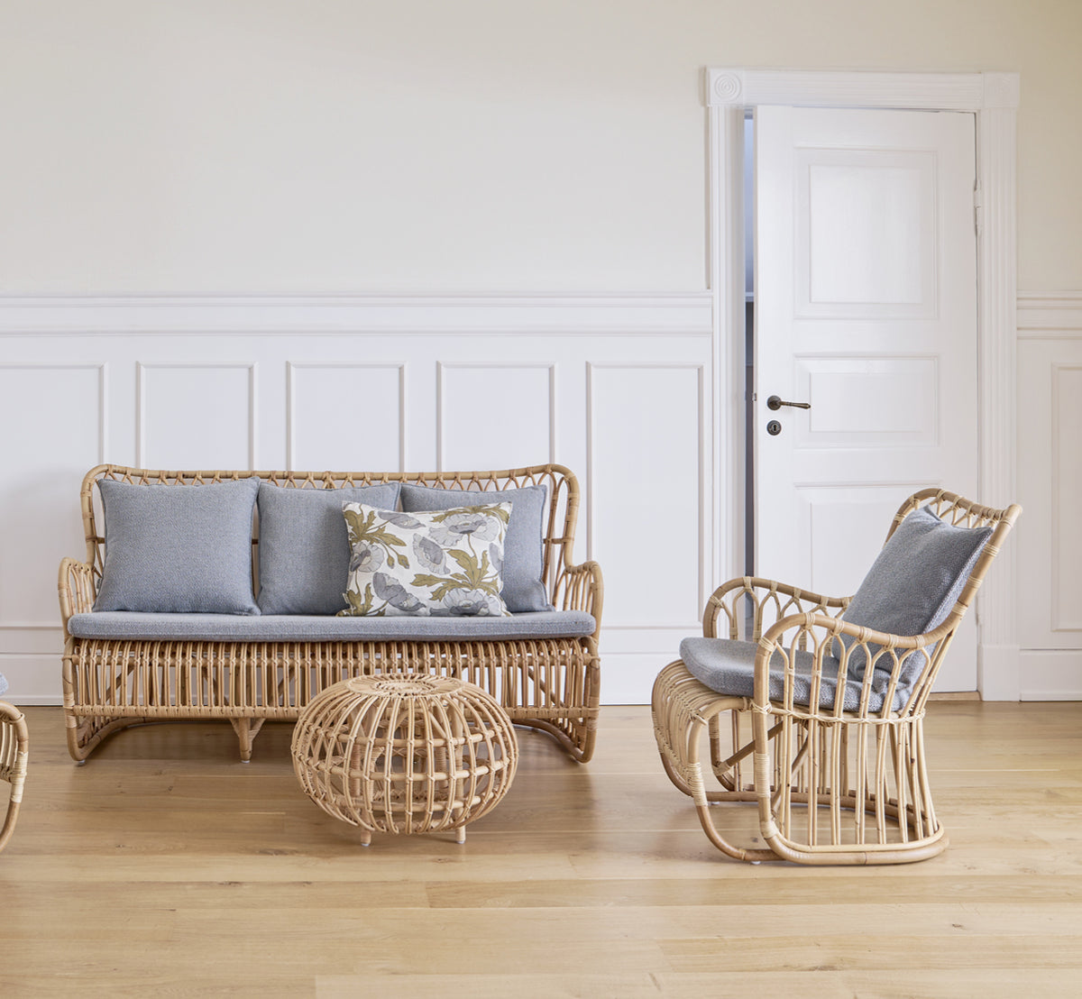 Esta es una imagen de estilo de vida de Tulip sofá en polished natural rattan. Combinada con un sillón lounge de ratán junto a una ventana soleada con cortinas.