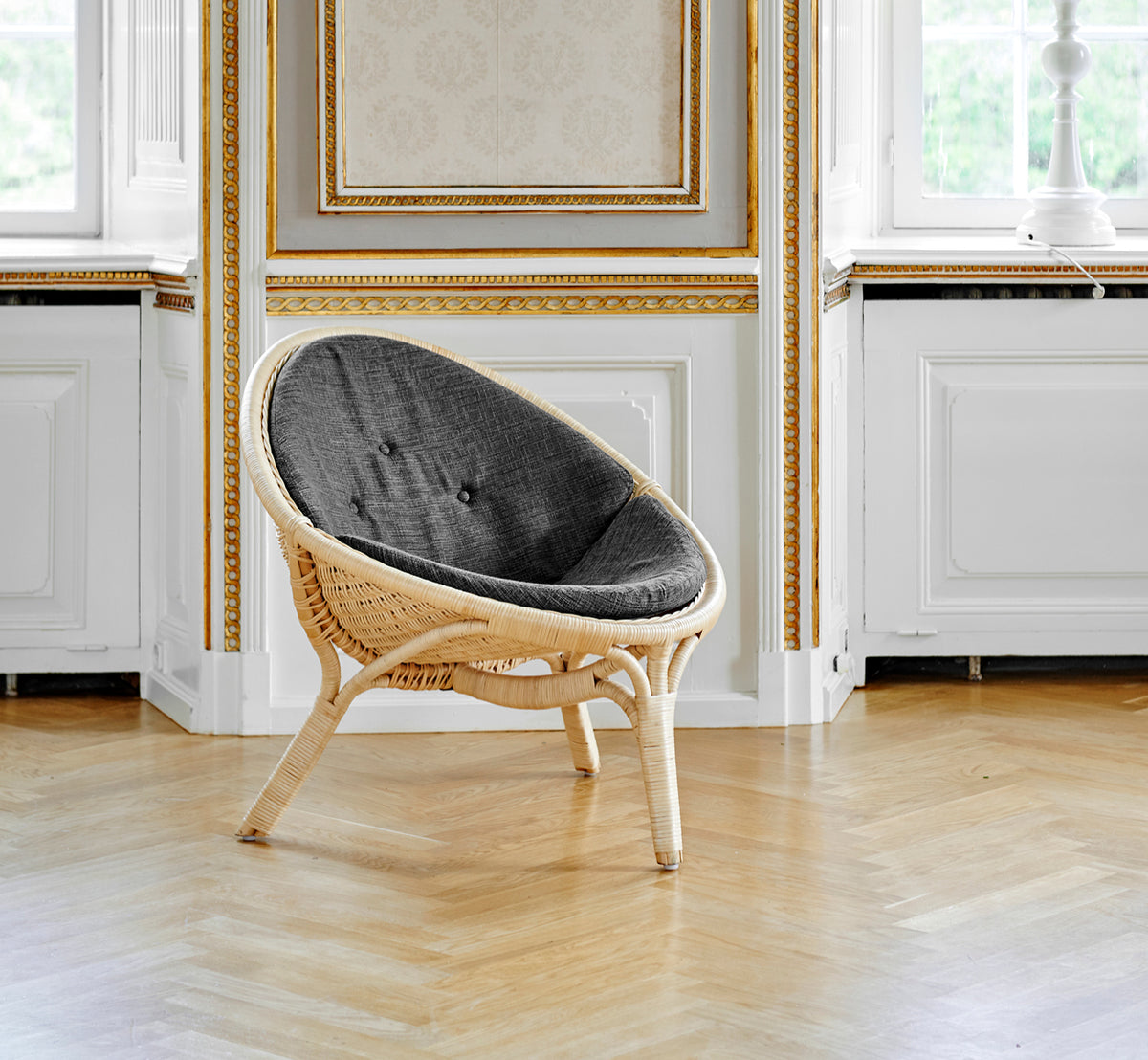 Esta es una imagen de estilo de vida de Rana sillón lounge en natural core weave rattan frame. De pie sola en una sala con paneles dorados y suelo de parqué.