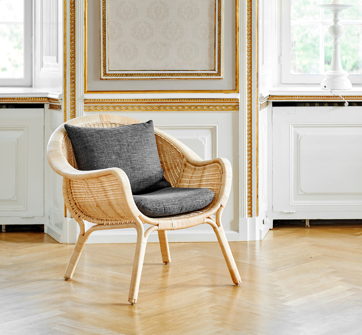Esta es una imagen de estilo de vida de Madame sillón lounge en natural core weave rattan frame. Elegante sala dorada con suelo de roble y cojín gris.