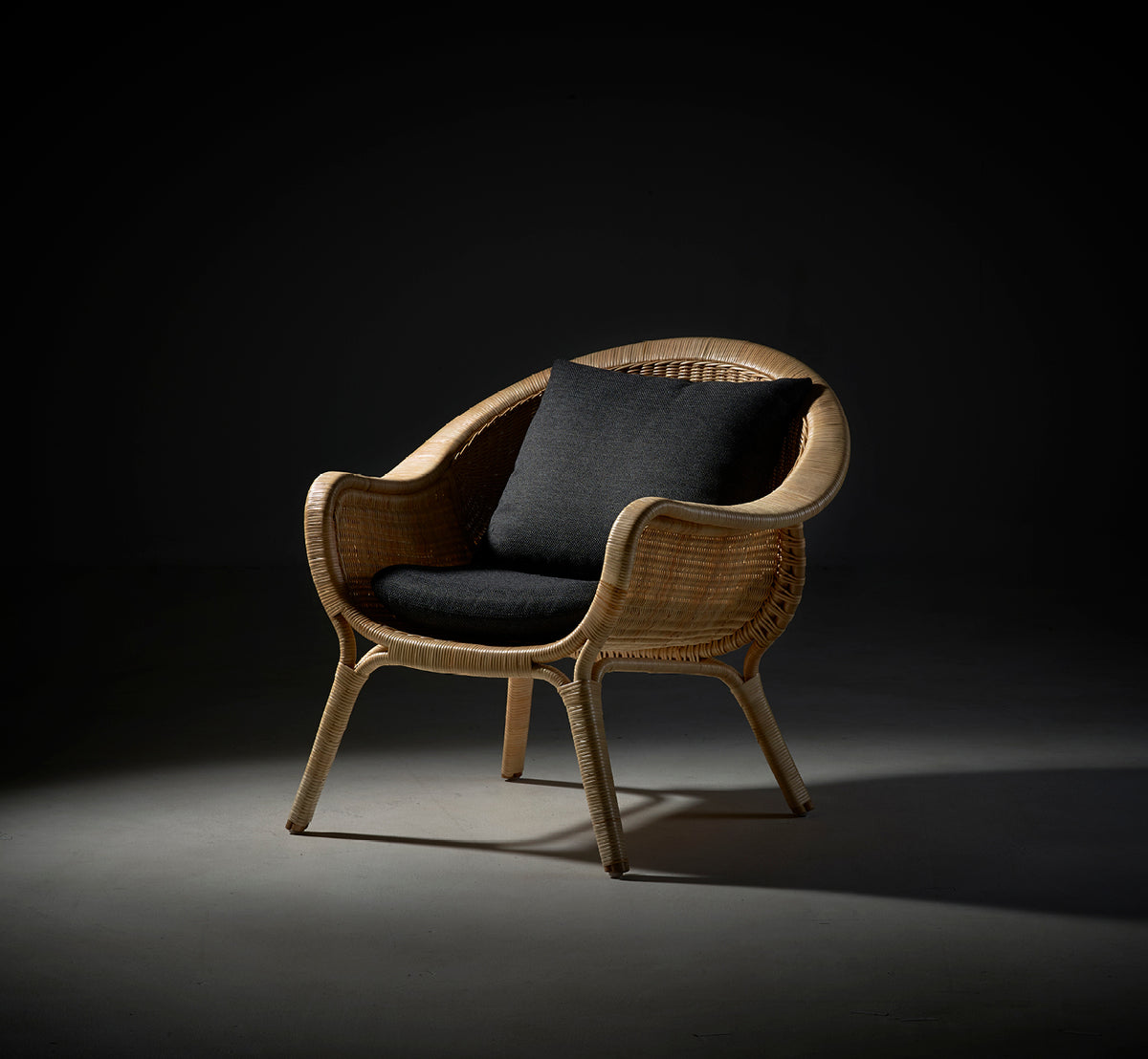 Esta es una imagen de estilo de vida de Madame sillón lounge en natural core weave rattan frame. Iluminación dramática de estudio con cojín oscuro sobre fondo gris.