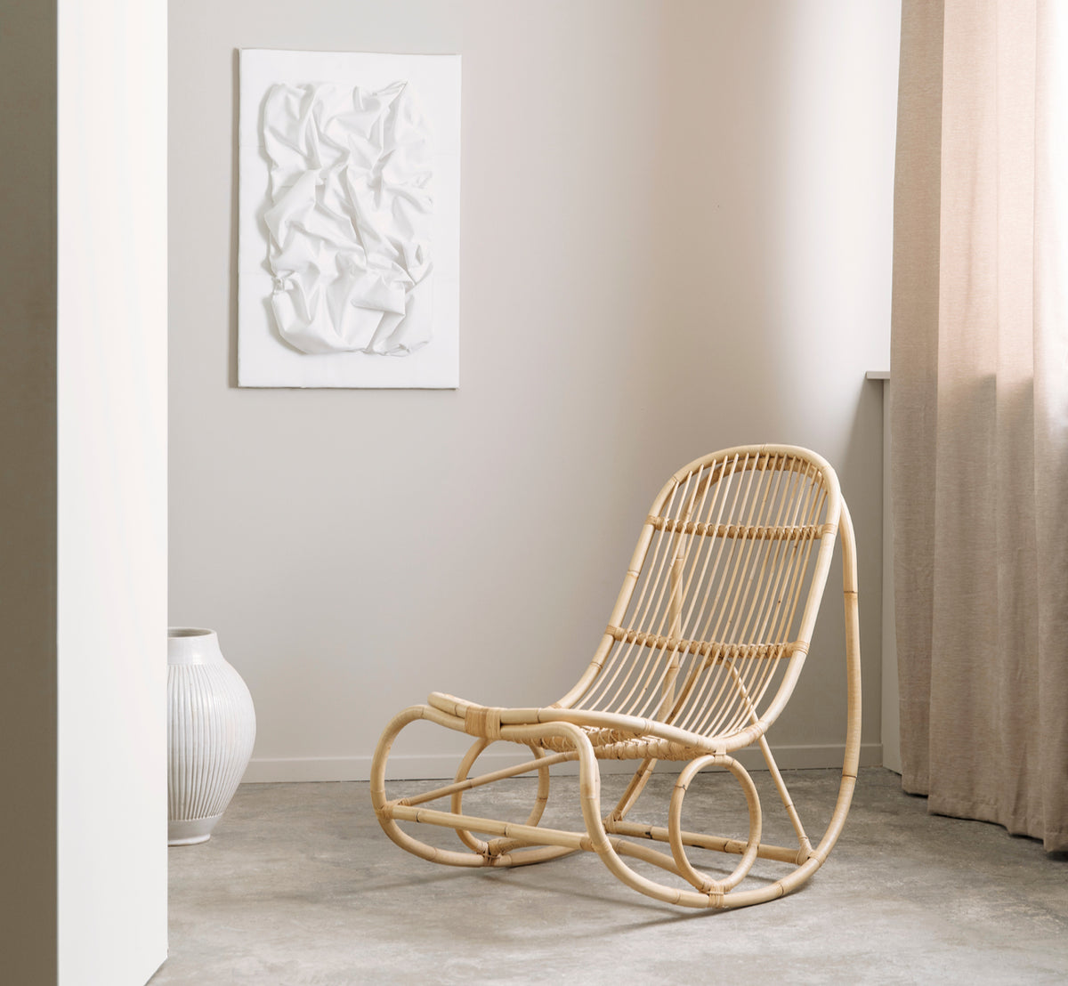 Esta es una imagen de estilo de vida de Nanny Rocking Chair i polished natural rattan. En un entorno de galería con otras piezas de ratán sobre pedestales.