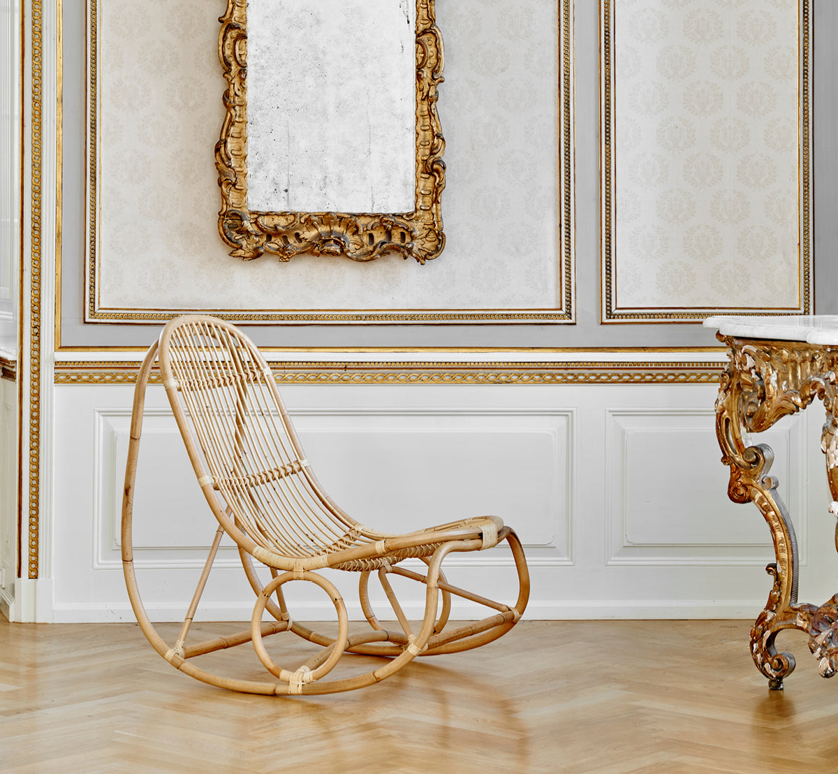 Esta es una imagen de estilo de vida de Nanny Rocking Chair i polished natural rattan. En una sala ornamentada dorada con espejo barroco y consola.