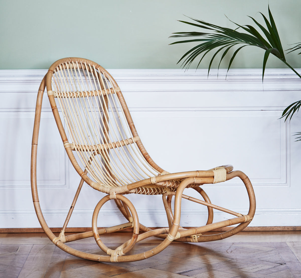 Esta es una imagen de estilo de vida de Nanny Rocking Chair i polished natural rattan. Junto a una pared verde salvia con paneles blancos y palmera.