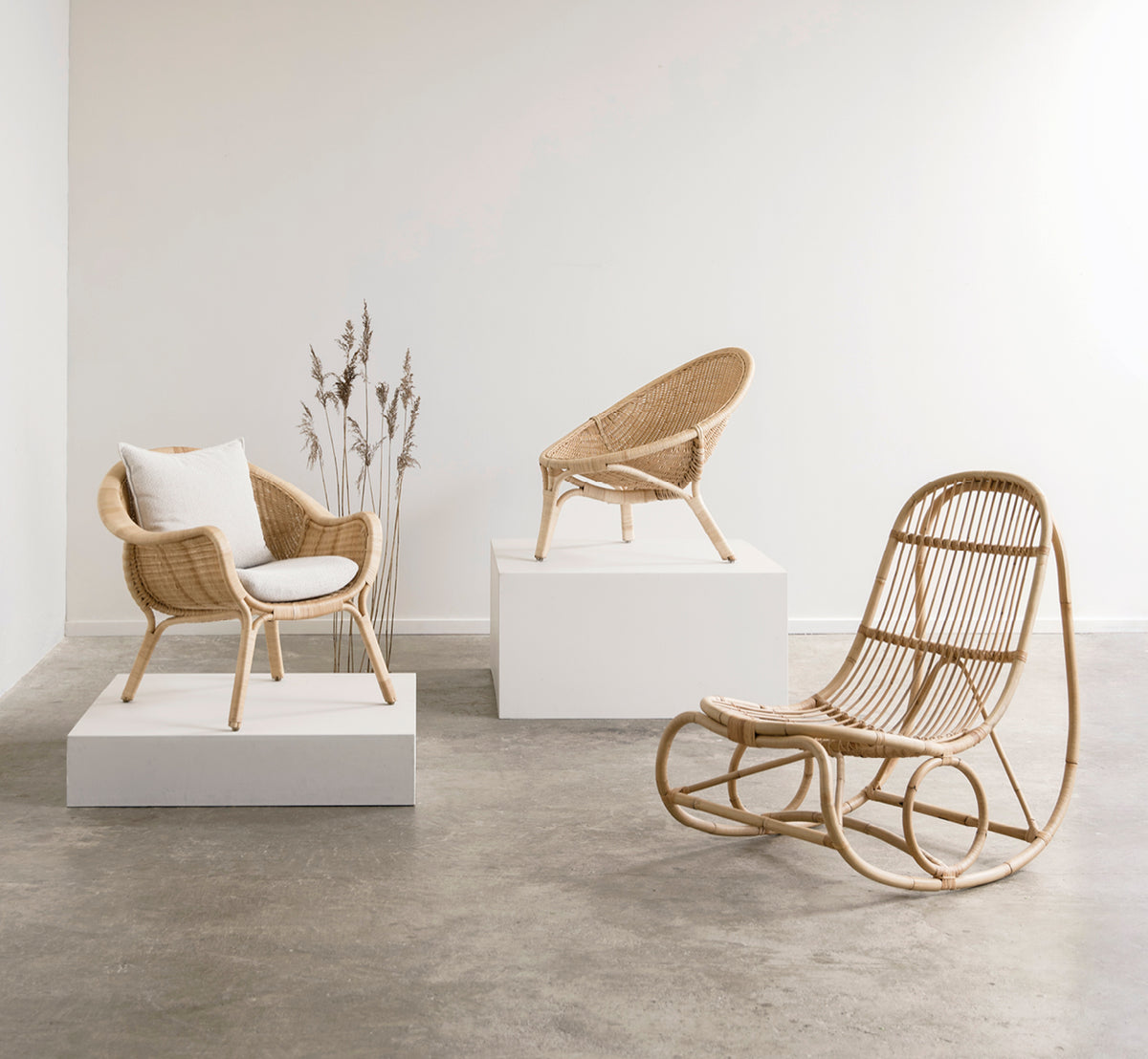 Esta es una imagen de estilo de vida de Nanny Rocking Chair i polished natural rattan. En una galería con sillas de ratán sobre pedestales blancos de exposición.