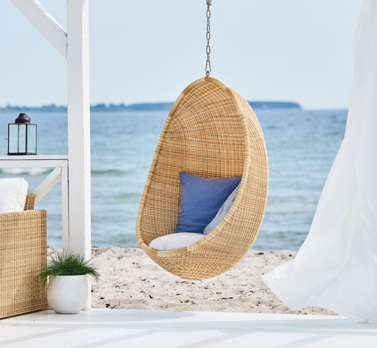 Esta es una imagen de estilo de vida de Hanging silla colgante exterior en natural core weave ArtFibre Aluminium, colgado de una pérgola blanca con.
