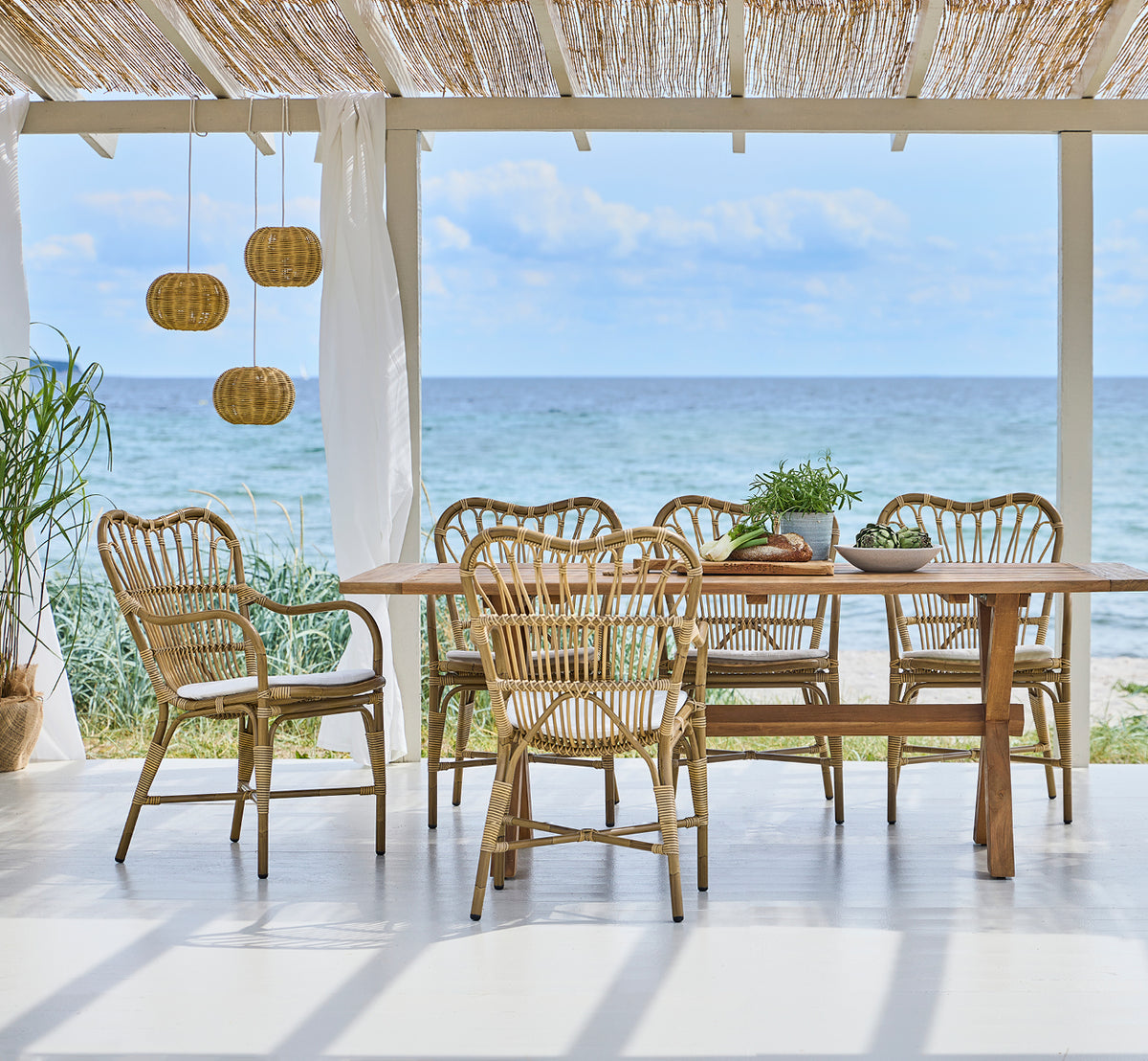 Esta es una imagen de estilo de vida de Margret sillón de comedor exterior en antique aluminium artfibre. Pabellón de playa con lámparas colgantes y vista al mar.