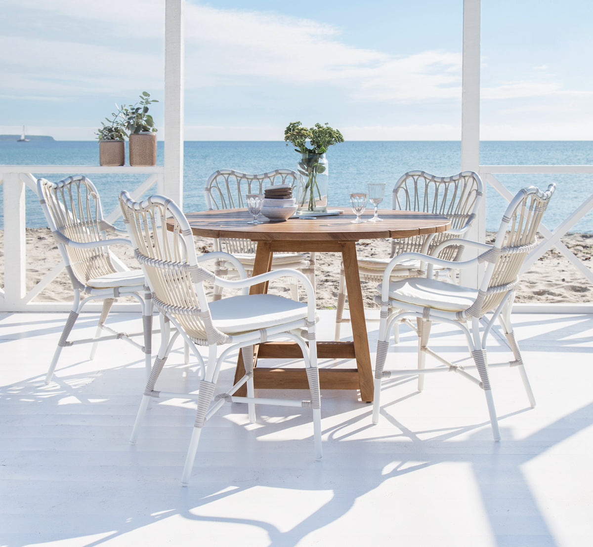 Esta es una imagen de estilo de vida de Margret sillón de comedor exterior en dove white aluminium artfibre. Terraza de playa con mesa de teca y vista al mar.