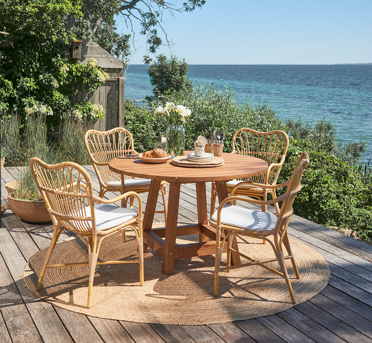 Esta es una imagen de estilo de vida de Margret sillón de comedor exterior en natural aluminium artfibre. Terraza junto al mar con mesa redonda de teca y plantas.