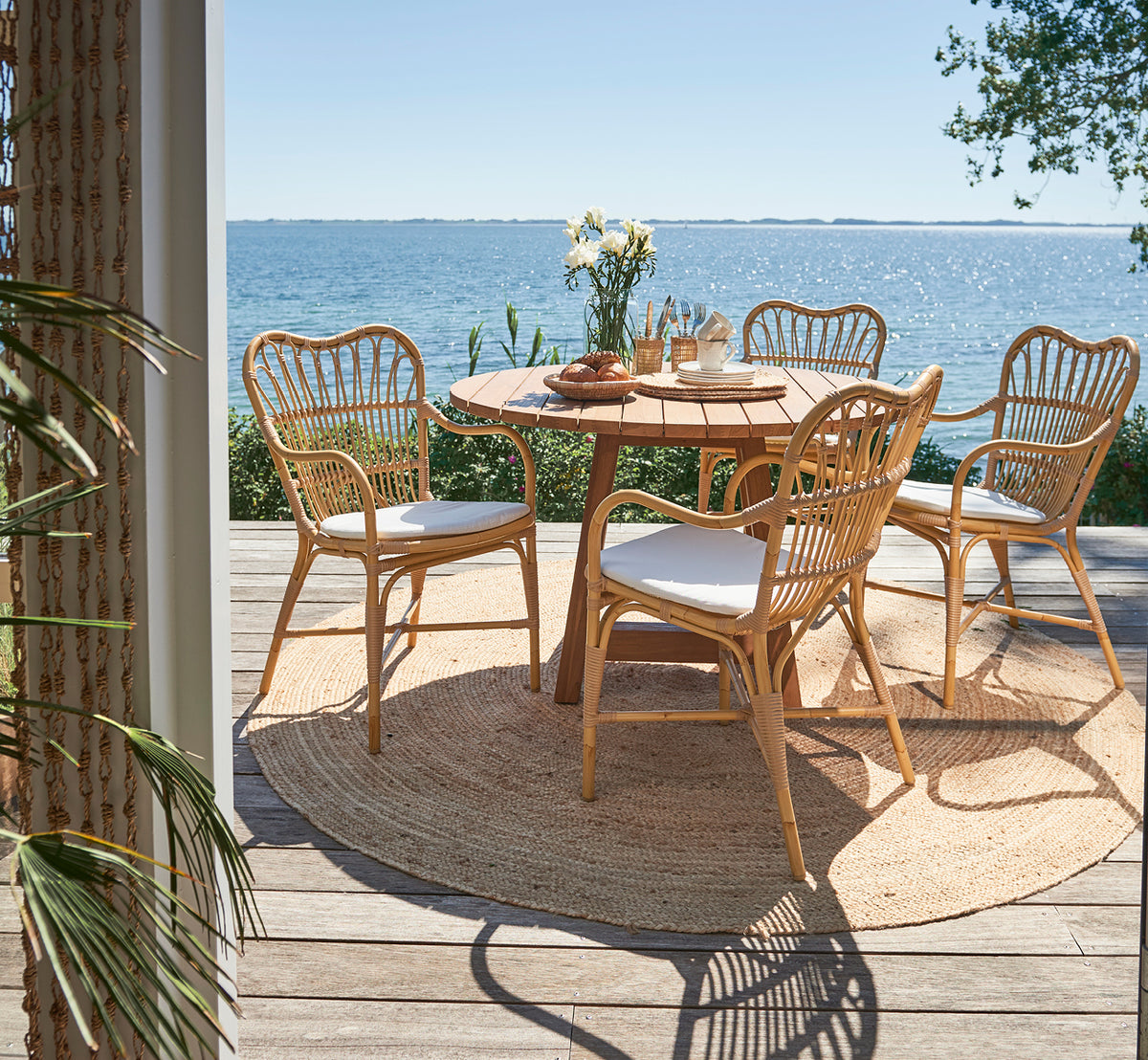 Esta es una imagen de estilo de vida de Margret sillón de comedor exterior en natural aluminium artfibre. Terraza junto al mar con mesa redonda de teca y vista al mar.