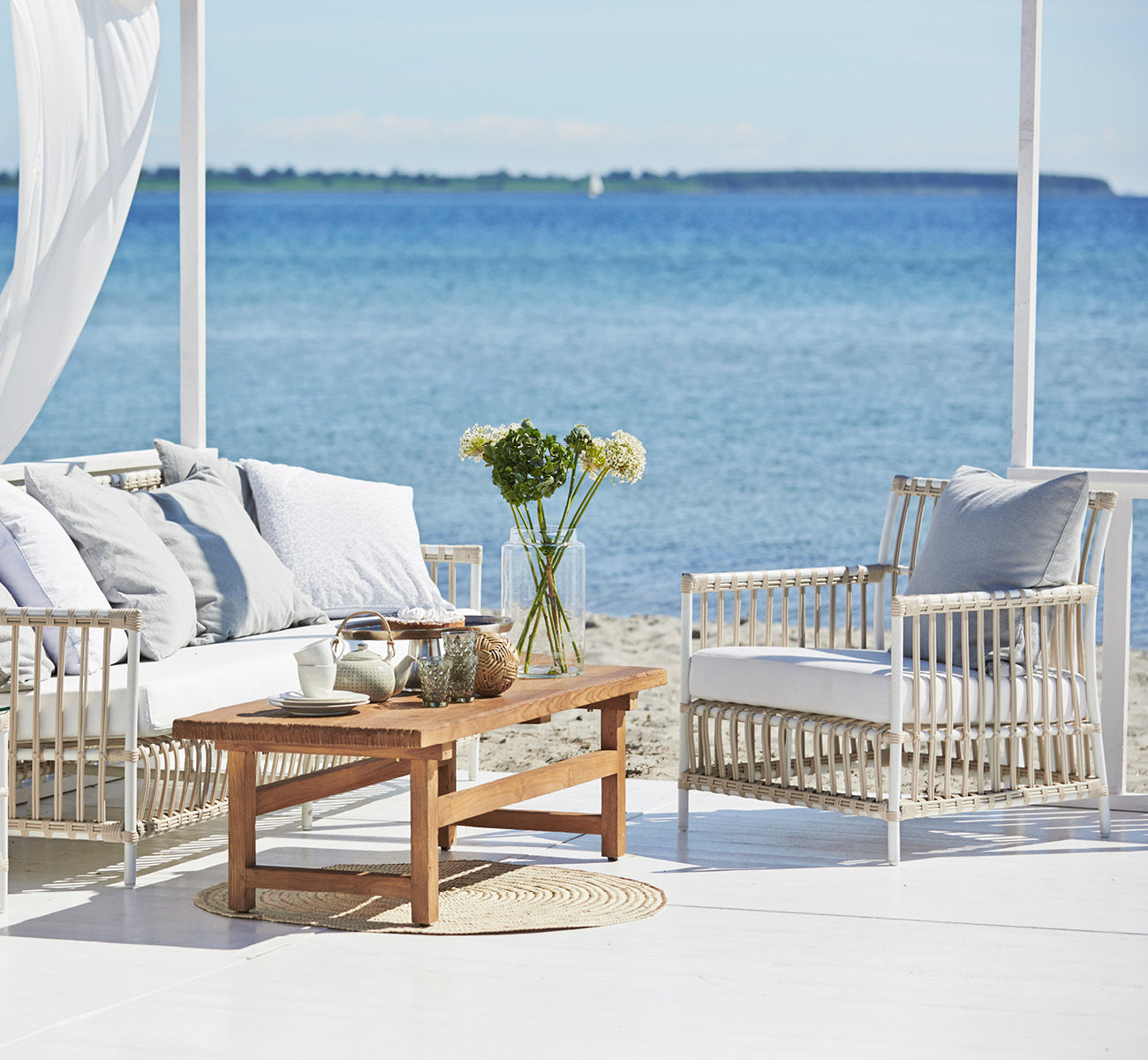 Esta es una imagen de estilo de vida de Caroline sillón lounge exterior en dove White Aluminium ArtFibre, en una terraza de madera junto al agua con.