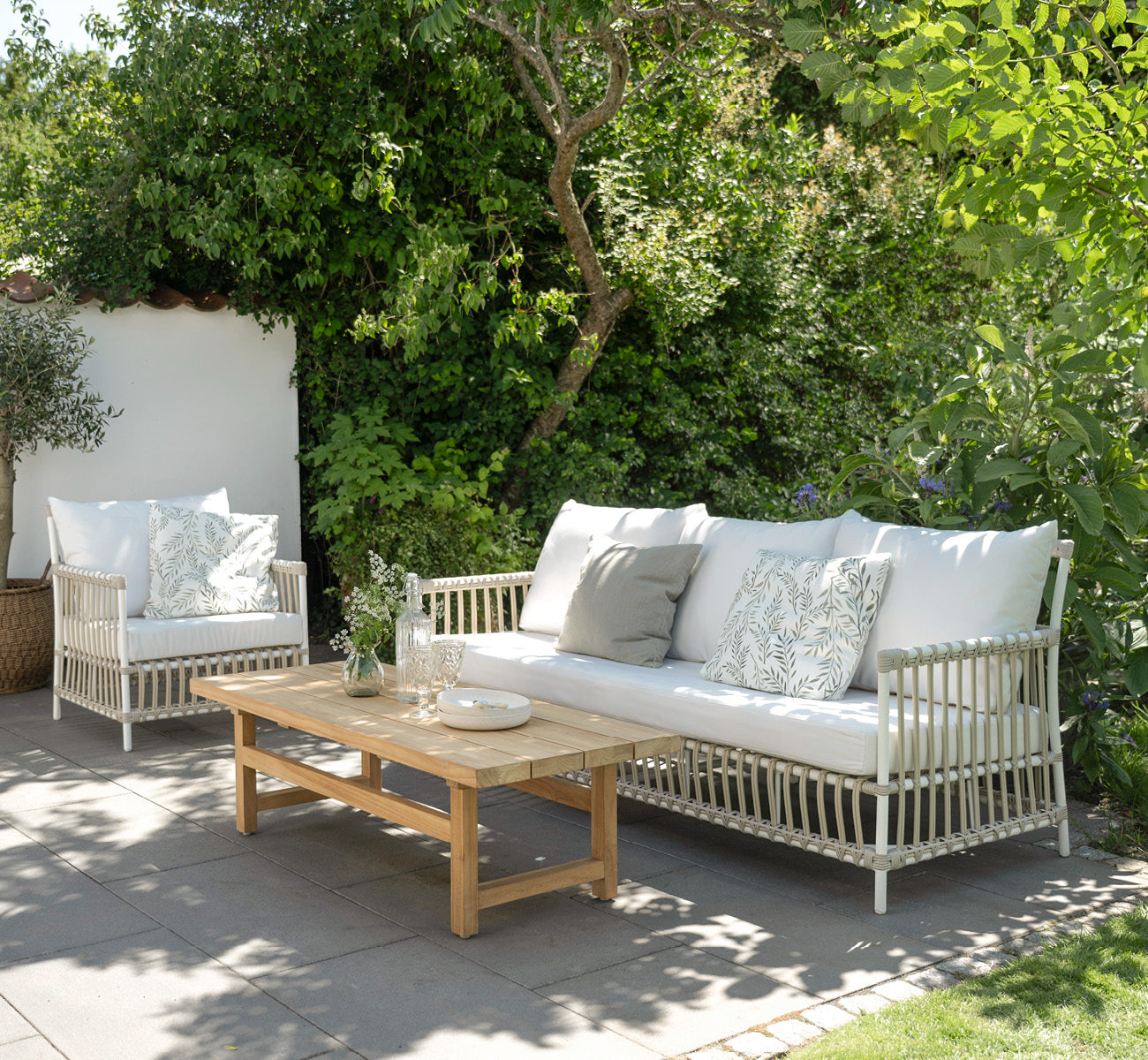 Esta es una imagen de estilo de vida de Caroline sillón lounge exterior en dove White Aluminium ArtFibre, en una terraza de madera junto al agua con.