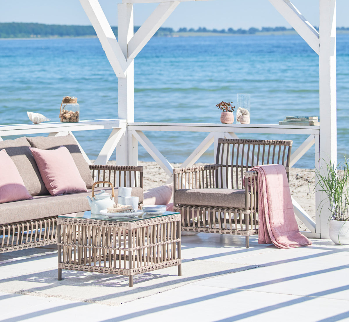 Esta es una imagen de estilo de vida de Caroline sillón lounge exterior en moccaccino Aluminium ArtFibre, en una terraza de madera junto al agua con.