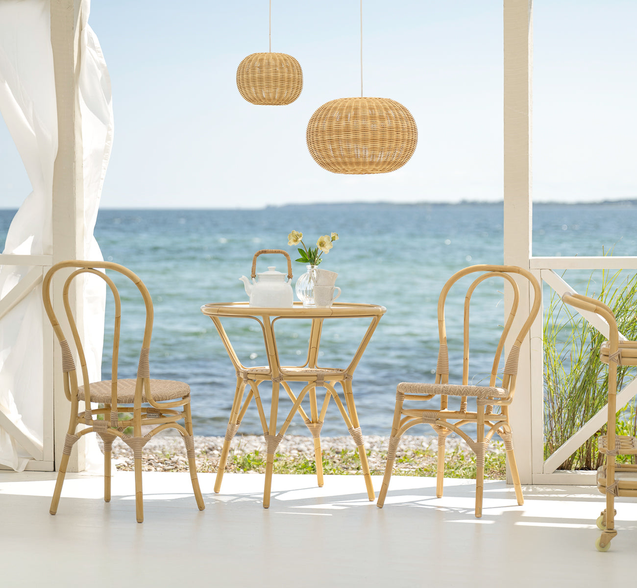 Esta es una imagen de estilo de vida de Lulu silla de comedor exterior en natural aluminium artfibre. En una terraza junto al mar con una mesa redonda y vista al océano.