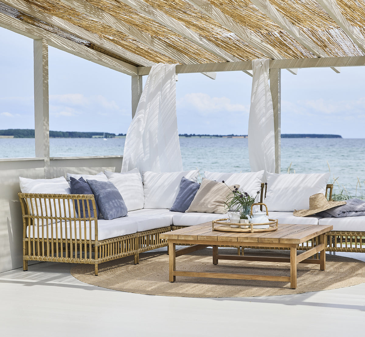Esta es una imagen de estilo de vida de Maggie Sofá Módulo Esquinero en antique aluminium artfibre. Pabellón de playa con conjunto de sofá y vista al mar.