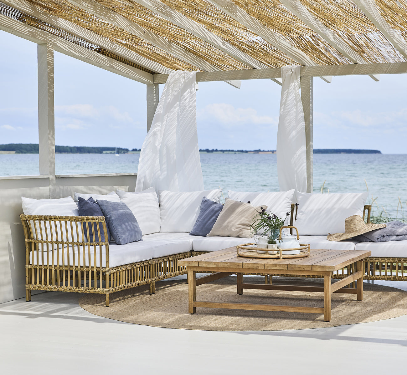 Esta es una imagen de estilo de vida de Maggie Sofá Módulo Esquinero en antique aluminium artfibre. Pabellón de playa con conjunto de sofá y vista al mar.