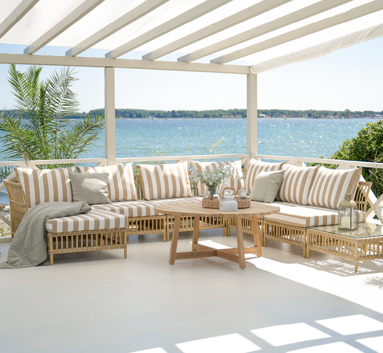 Esta es una imagen de estilo de vida de Maggie Sofá Módulo Esquinero en natural aluminium artfibre. Terraza cubierta con cojines a rayas y vista al mar.