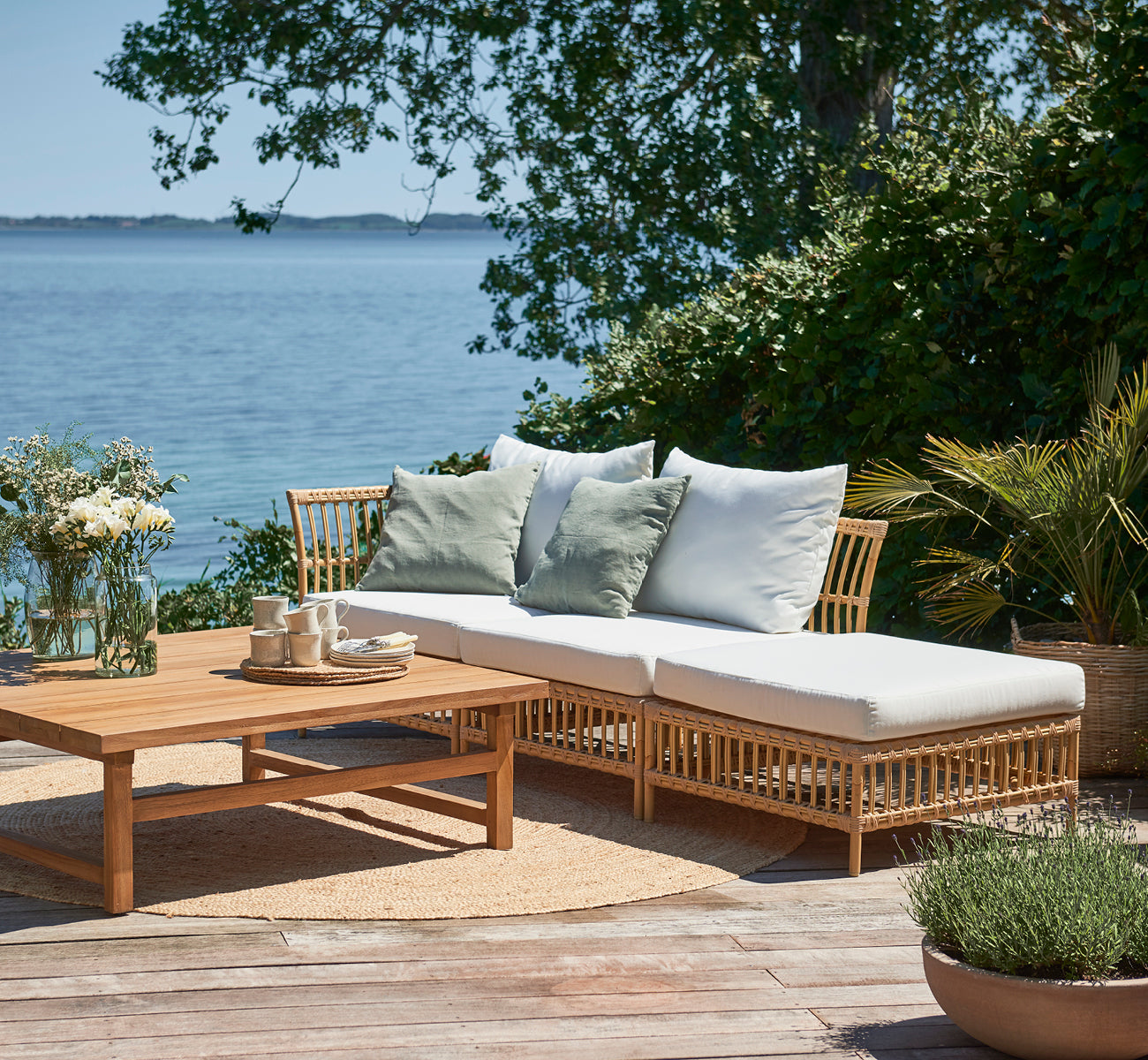 Esta es una imagen de estilo de vida de Maggie Sofá Módulo Esquinero en natural aluminium artfibre. Terraza junto al mar con mesa de centro y cojines verdes.