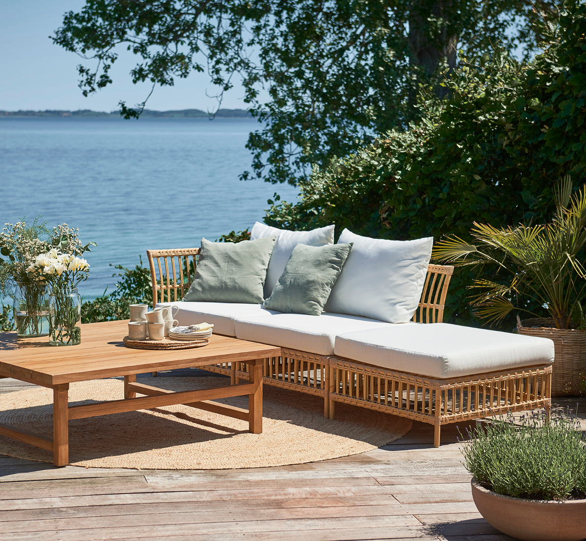 Esta es una imagen de estilo de vida de Maggie Sofá Módulo Central en natural aluminium artfibre. Terraza junto al mar con cojines, plantas y vista al mar.