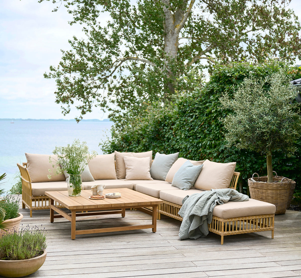Esta es una imagen de estilo de vida de Maggie Sofá Módulo Central en natural aluminium artfibre. Terraza junto al mar con sofá modular y vegetación alrededor.