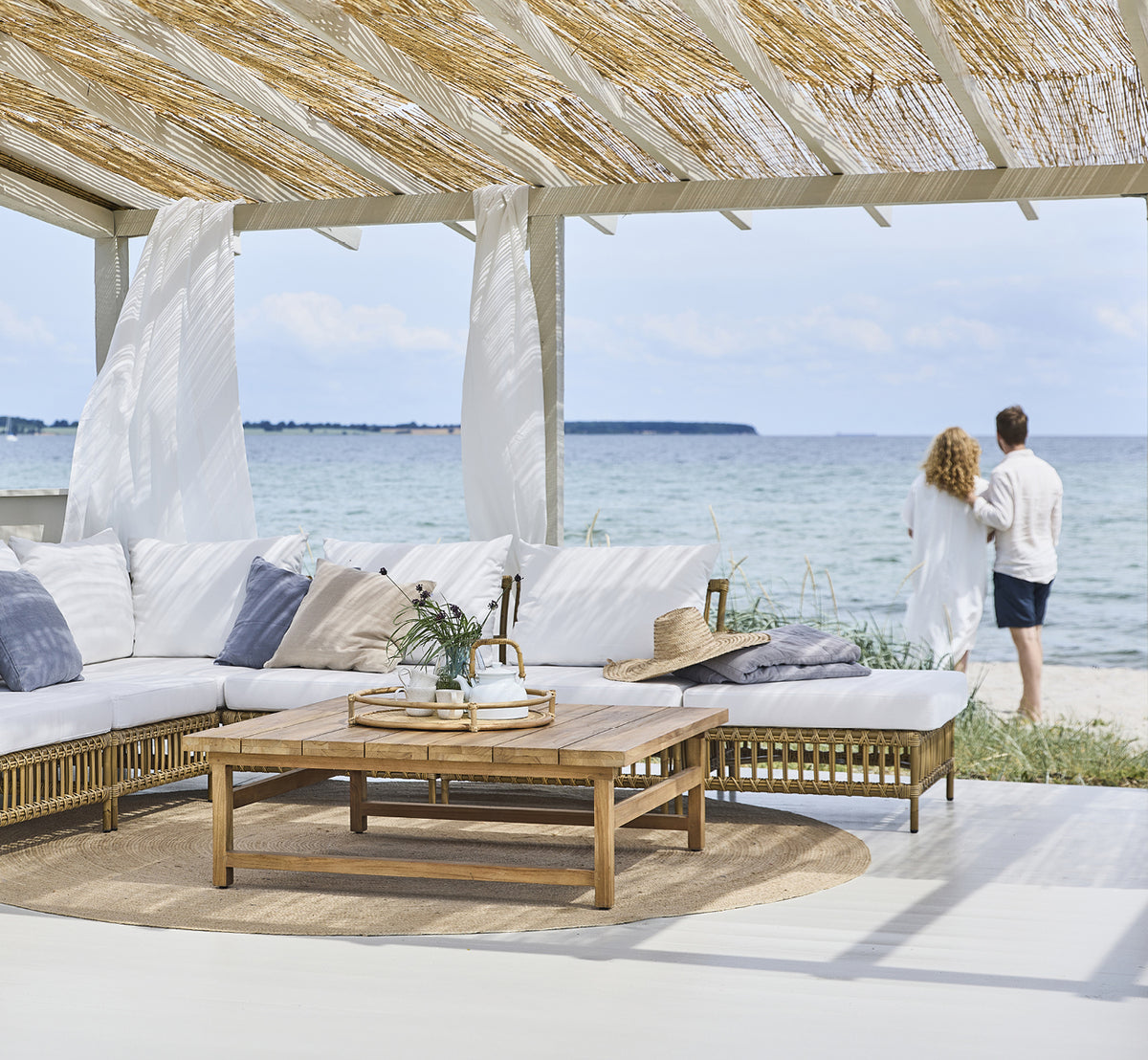 Esta es una imagen de estilo de vida de Maggie Sofá Módulo Puf c. Cristal en antique aluminium artfibre. Pabellón de playa con pareja y vista al mar.