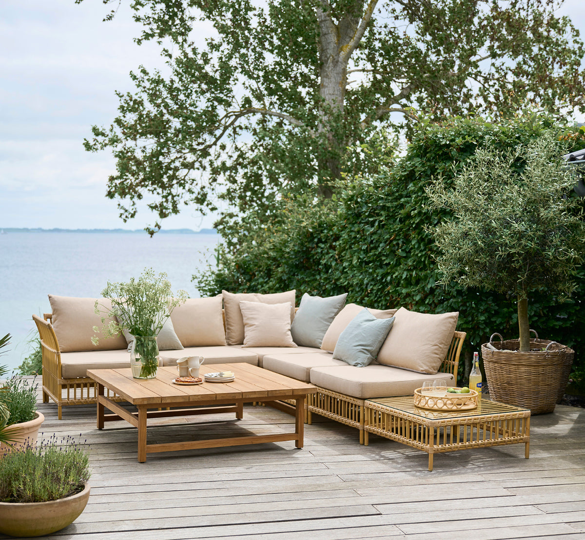 Esta es una imagen de estilo de vida de Maggie Sofá Módulo Puf c. Cristal en natural aluminium artfibre. Terraza junto al mar con conjunto de sofá y vegetación.