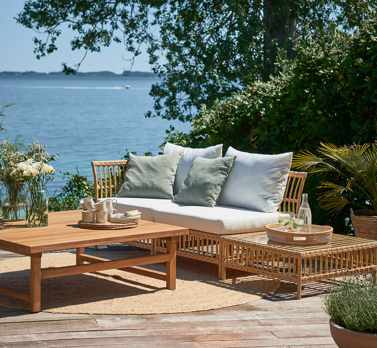 Esta es una imagen de estilo de vida de Maggie Sofá Módulo Puf c. Cristal en natural aluminium artfibre. Terraza junto al mar con mesa de centro y cojines verdes.