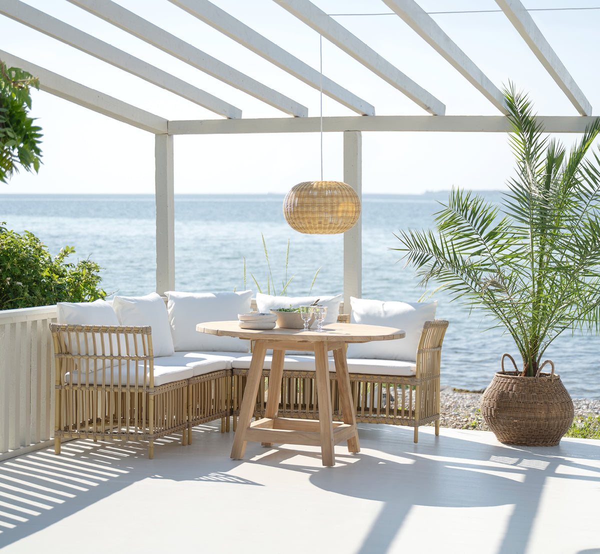 Esta es una imagen de estilo de vida de Meghan Exterior Dining Bench Corner Module i natural aluminium artfibre. Con mesa redonda junto al mar.