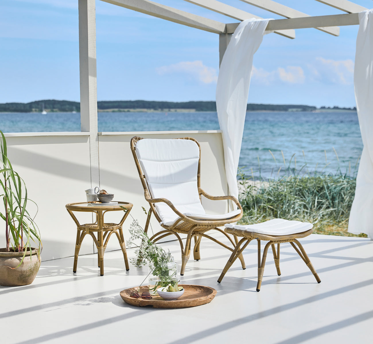 Esta es una imagen de estilo de vida de Monet Exterior Lounge Chair i antique aluminium artfibre. Con reposapiés en la terraza de un pabellón junto al mar.