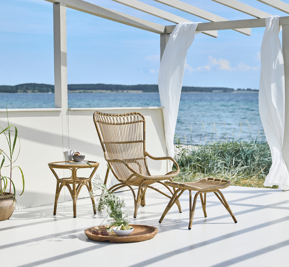 Esta es una imagen de estilo de vida de Monet Exterior Lounge Chair i antique aluminium artfibre. Con reposapiés en una terraza de playa con vista al mar.