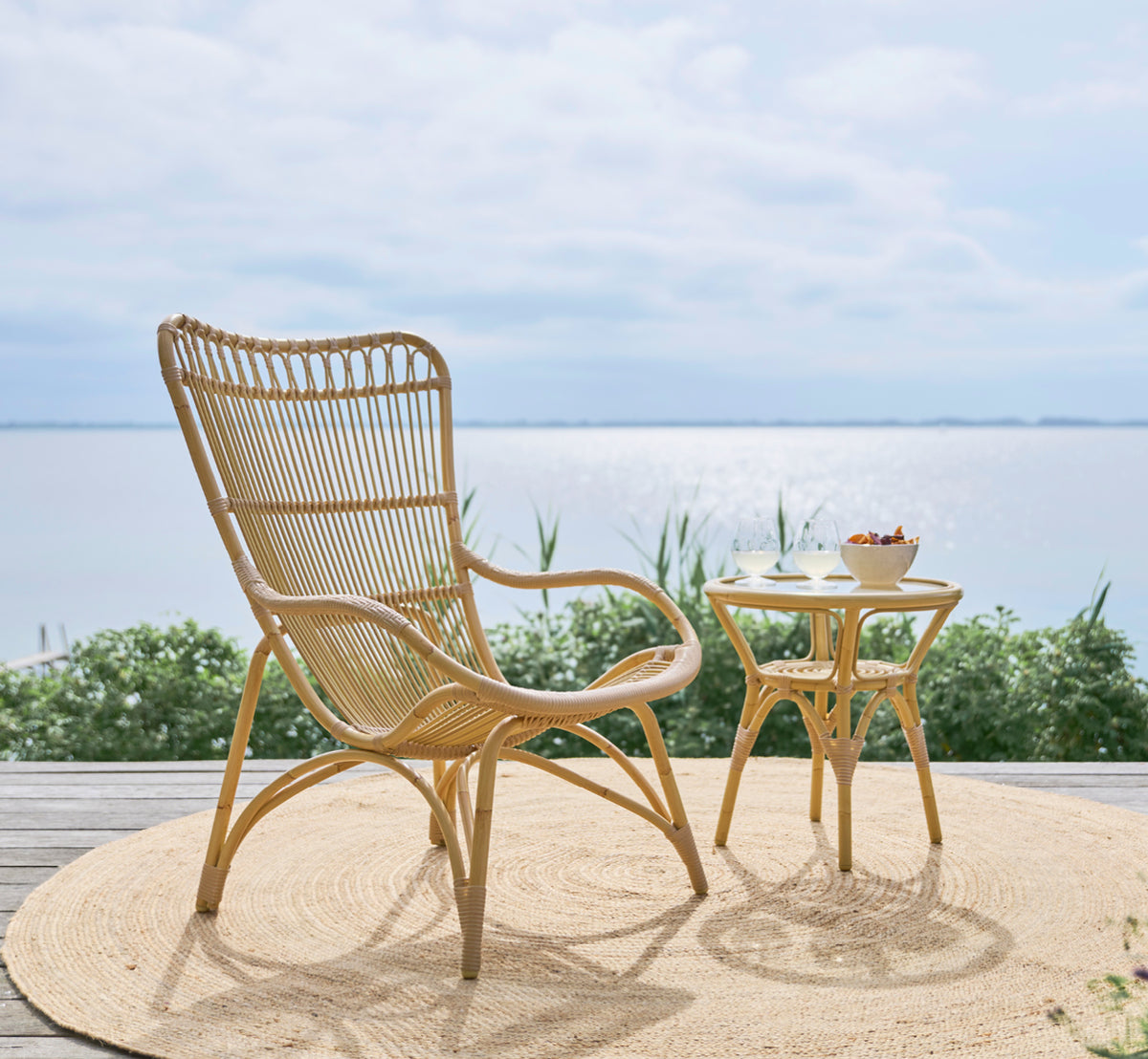 Esta es una imagen de estilo de vida de Monet Exterior Lounge Chair i natural aluminium artfibre. En una terraza de madera con alfombra de yute y vista al mar.