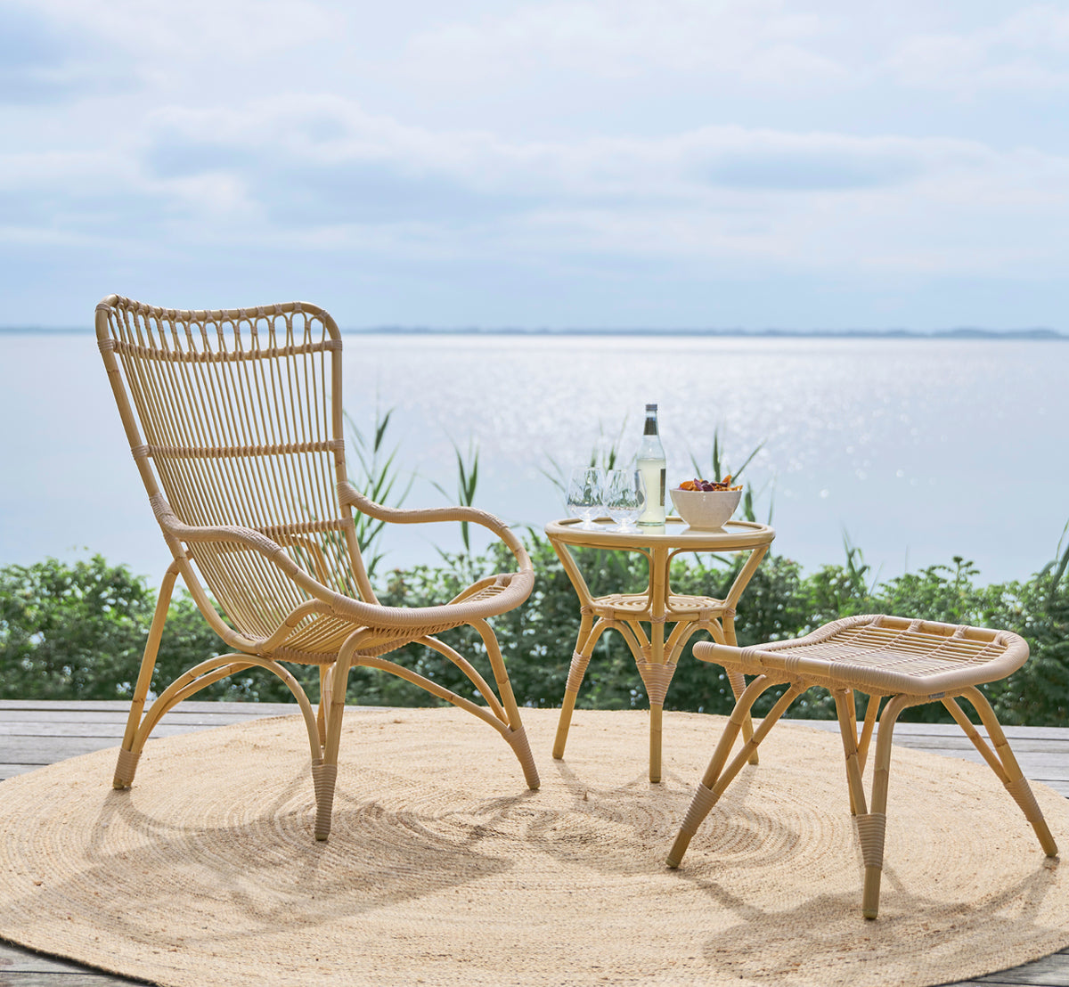Esta es una imagen de estilo de vida de Monet Exterior Lounge Chair i natural aluminium artfibre. Con reposapiés en una terraza con vistas al mar.